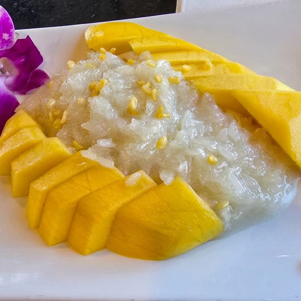 Mango sticky rice topped with toasted mung beans, arranged on a white plate with a purple orchid garnish.
