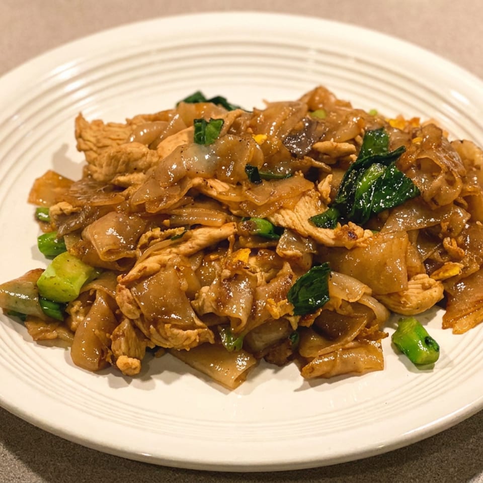 Stir-fried wide rice noodles with sliced meat, leafy greens, and vegetables on a white plate.