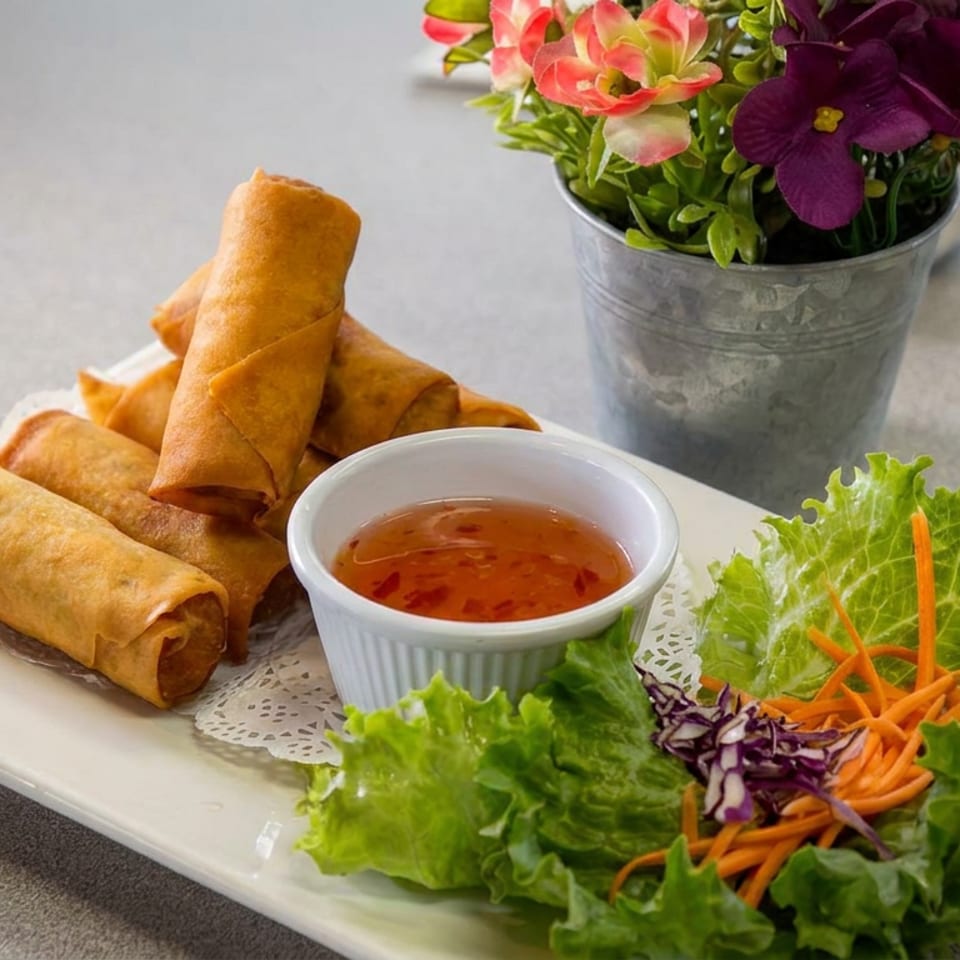 Crispy spring rolls with sweet chili dipping sauce, lettuce, shredded carrots, and purple cabbage on a white plate.