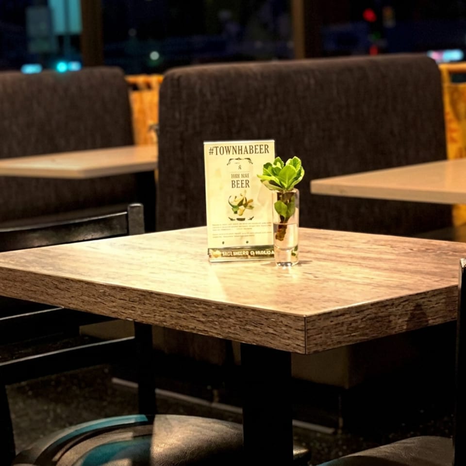 Warmly lit restaurant dining area with a wooden table, black chairs, booth seating, a beer promotion card, and a small potted plant.